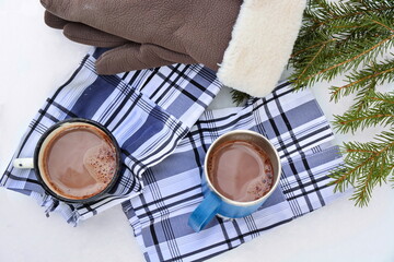 Pair of iron mugs with hot chocolate or cocoa on a checkered fabric next to fir branches and a pair of gloves in the snow. Outdoor picnic in winter. Top view