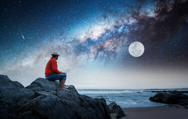 Fototapeta na wymiar person sitting on a rock with a laptop on the beach staring the moon and Milky Way at night