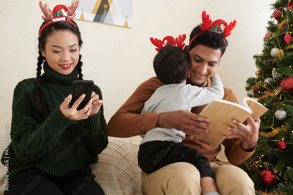 Poster Smiling young woman ordering Christmas presents online when husband reading book to little son