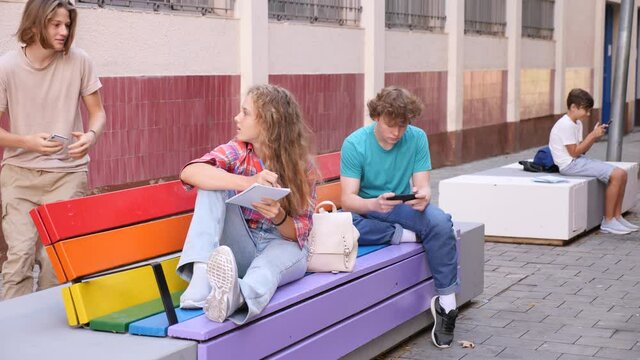 Teenager girl and young boy sitting on bench. Girl doing homework and talking with her friend, young boy using smartphone.