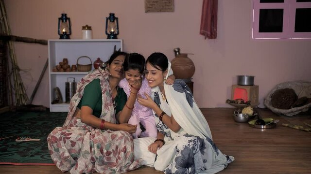 A little child from a village in India hugging her mother and aunt sitting together - Family in village . A sweet girl from a rural household sitting and playing with her toys in the kitchen at hom...