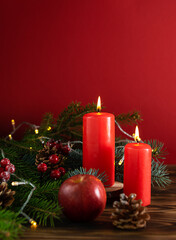 Red candles on a background of green fir branches, cones and gifts. Red berries and apples in the background. Dark red background, space for text