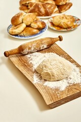 Wooden cutting board with raw dough for pies from wheat flour and vintage rolling pin on white table near fresh homemade sweet and meat pies