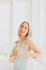 Young happy woman stands by the window glass of refreshing drink with lemon and mint. Healthy lifestyle, rejuvenation