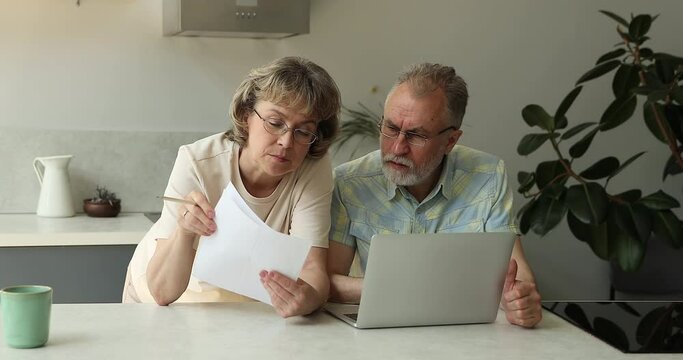 Serious senior adult couple focused on paperwork at home office. Family business owners read paper draft loan contract credit agreement check terms discuss conditions fill in application form online