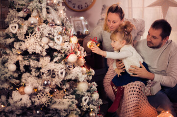 Merry Christmas and Happy Holidays! Dad, Mom and daughter decorate the Christmas tree indoors. The morning before Xmas. Portrait loving family close up. - 475972471