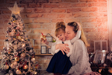 Merry Christmas and Happy Holidays! Mom and daughter decorate the Christmas tree indoors. The morning before Xmas. Portrait loving family close up. - 475972495