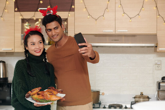 Happy young husband and wife with reindeer antlers headband taking selfie and showing grilled chicken cooked for Christmas dinner