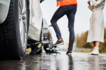 Close up of automobile on rainy road with female driver and roadside assistance worker on blurred background. Male auto mechanic repairing woman car on the street. Focus on care wheel.