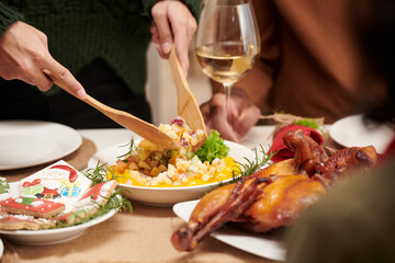 Hands of woman using wooden spoon when putting salad from main dish in her plate at family dinner