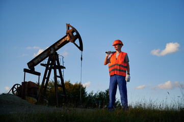 Successful technician in uniform and helmet enjoying occupation job at derrick pump jack during shift at oil station, male inspector with instrument smiling during offshore maintenance for mine