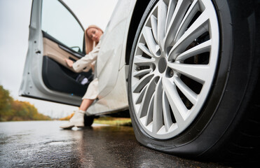 Close up view of rear semi-lowered wheel of white automobile parked on wet road. Blonde female driver stopped her car on edge of road and looking at punctured tire of back wheel. - Powered by Adobe