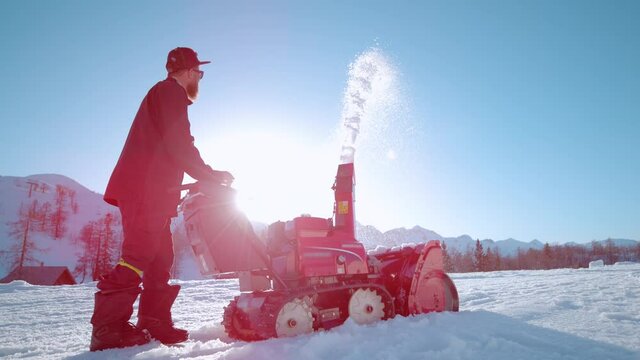SLOW MOTION, LOW ANGLE, LENS FLARE: Bright sunbeams shine on a bearded male shaper using a snow blower to create a kicker out of fresh snow. Machinery operator blows snow into air on sunny winter day.