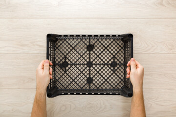 Young adult man hands holding empty black plastic crate on light wooden table background. Closeup. Point of view shot. Grating box for different products delivery or storage. Top down view.