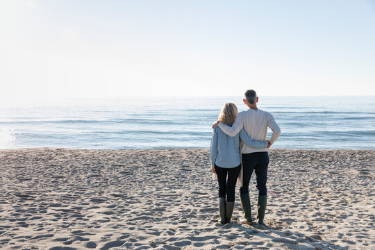 Man With Arm Around Woman Looking At Sea Standing On Sand