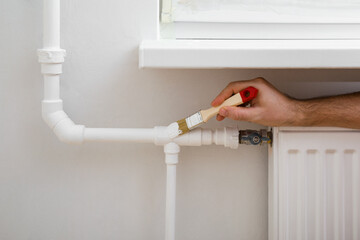 Young adult man hands using brush and painting heating pipe white color. Closeup. Repair work of...