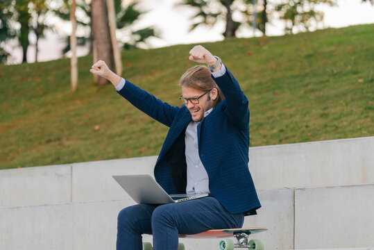 Happy businessman with laptop cheering sitting on longboard on steps