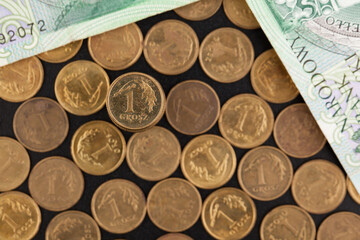 In the foreground a one penny coin, in the background blurred, scattered coins and two banknotes, lying on a dark background.