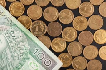 In the foreground a one penny coin, in the background blurred, scattered coins and a 100 zloty bill, lying on a dark background.