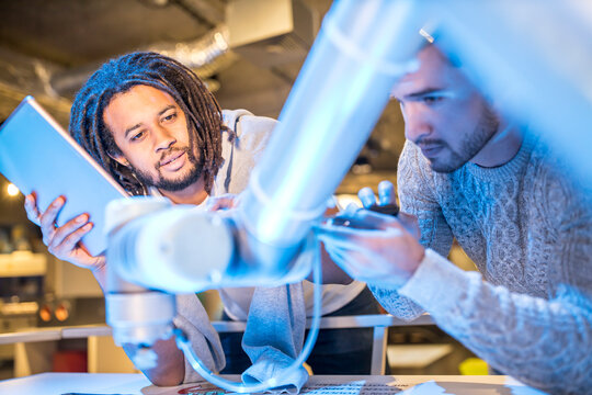 Young technician with tablet PC discussing with colleague repairing robotic arm at workshop