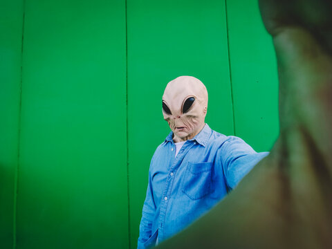 Standing alien mask face taking selfie picture against green background wall. Copy space for comic funny advertising. Ufo extraterrestrial incasual denim clothes use mobile phone