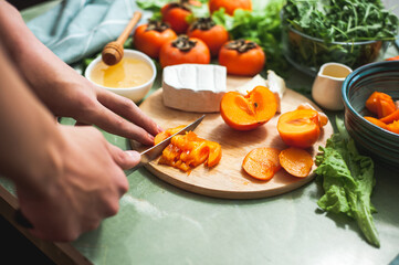 Ingredients and process of making a vitamin winter salad with persimmons, tangerines and cheese Female or teenage hand cuts persimmon on a round wooden board Vegan lunch preparation. Selective focus