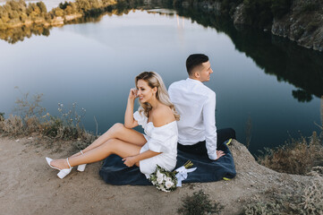 A stylish young groom and a beautiful blonde bride in a white dress are sitting on a rug on a cliff, with their backs to each other, against the backdrop of rocks and a river. Wedding photography.