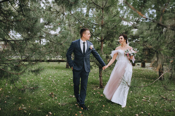 Stylish groom in a gray suit and a beautiful bride in a white dress are walking through the woods, holding hands on the green grass in nature. Wedding portrait of the newlyweds.