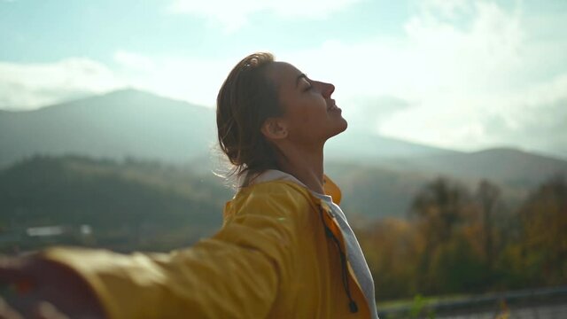 slow motion portrait Young woman wearing a yellow jacket raising her arms into blue sky and enjoying view above clouds on top of mountain.