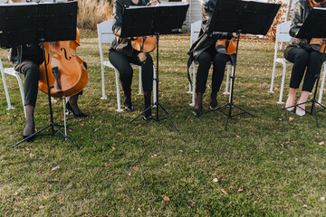 Girls musicians sit on white chairs outdoors in the park, playing the violin, cello, double bass.