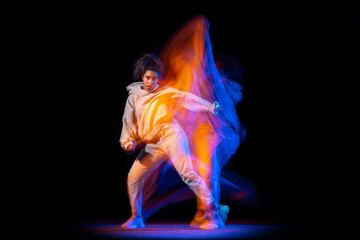 Sportive young girl in white costume dancing hip-hop dance isolated on dark background at dance hall in neon mixed light.