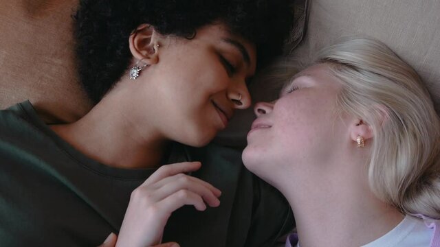 Young mixed lesbian kisses hug laying in bed and smile, shot from top. Lesbian couple hugging. Young couple happily talk. High quality 4k footage