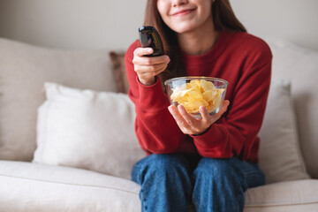 Closeup image of a woman eating potato chips while searching channel with remote control to watch tv at home