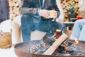 couple of young woman and man having fun, tenderness during teatime near fire in courtyard of suburban house in winter, concept of Christmas and New Year vacation on farm, family love and support