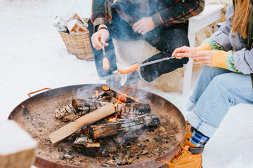 couple of young woman and man having fun and fry sausages on fire in courtyard of suburban house in winter, concept of Christmas and New Year vacation on farm, family love and support, winter fun