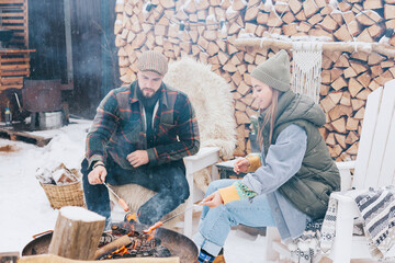 couple of young woman and man having fun and fry sausages on fire in courtyard of suburban house in winter, concept of Christmas and New Year vacation on farm, family love and support, winter fun