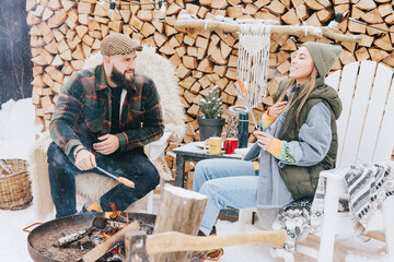 couple of young woman and man having fun and fry sausages on fire in courtyard of suburban house in winter, concept of Christmas and New Year vacation on farm, family love and support, winter fun