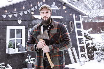 young handsome brutal bearded man with axe in hands, chop and gathering firewood in courtyard of suburban house in winter, concept of Christmas and New Year vacation on farm, family love and support