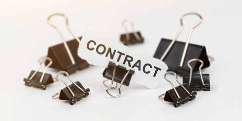 On a white table there are office clamps, in one clip there is paper with the inscription - CONTRACT