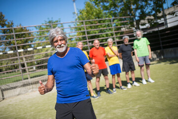 Portrait of funny senior man on football field. Team leader with gray hair in sport clothes standing, showing thumbs up, teammates in background. Football, sport, leisure concept