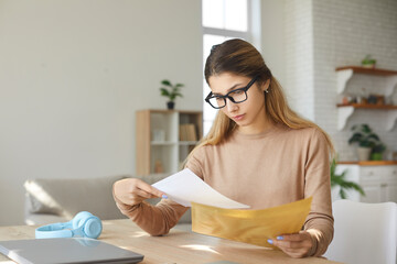 Anxious young woman at home taking letter from envelope reads bad negative news. Girl sitting at table reads notice from bank about debt on loan, about dismissal from work or notice of eviction. - obrazy, fototapety, plakaty