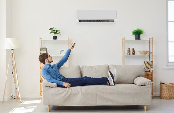 Happy man resting on couch under air conditioner enjoys fresh air. Man with remote control from air conditioner makes comfortable temperature for himself. Concept of climate system in modern house.