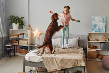 Full length portrait of cute blonde girl jumping with dog on bed, copy space