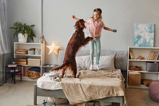 Full Length Portrait Of Cute Blonde Girl Jumping With Dog On Bed, Copy Space