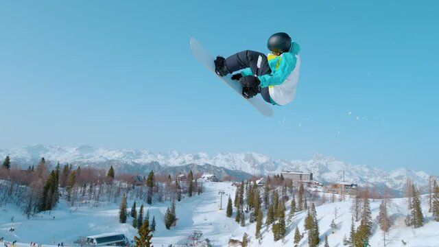 SLOW MOTION: Male snowboarder soars through the air and does a breathtaking trick. Spectacular shot of a snowboarding pro doing a rotating grab stunt while training in Vogel ski resort snowpark.
