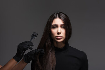 hairstylist holding brush near sad woman with brown hair isolated on dark grey - obrazy, fototapety, plakaty