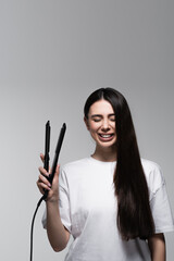 happy brunette woman holding hair straightener isolated on grey