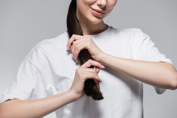 cropped view of cheerful woman in white t-shirt touching brown hair isolated on grey