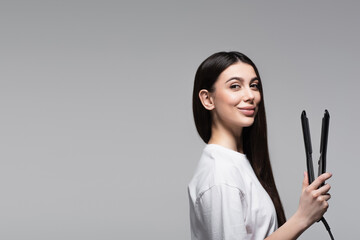 cheerful brunette woman holding hair straightener isolated on grey
