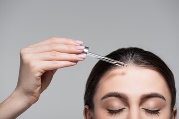 cropped view of young woman holding pipette with oil near shiny hair isolated on grey